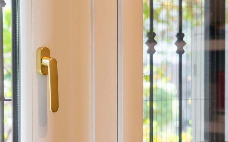 Mâner auriu pe o ușă de balcon din PVC albă, cu grilaj forjat și verdeață în spate